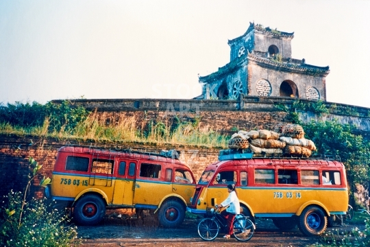 French colonial era buses in Vietnam - Iconic historic red yellow Renault bus model (probably a _qt_1000kg’) in front of the wall and gate tower of the Imperial City of Hue - old vintage low resolution photo